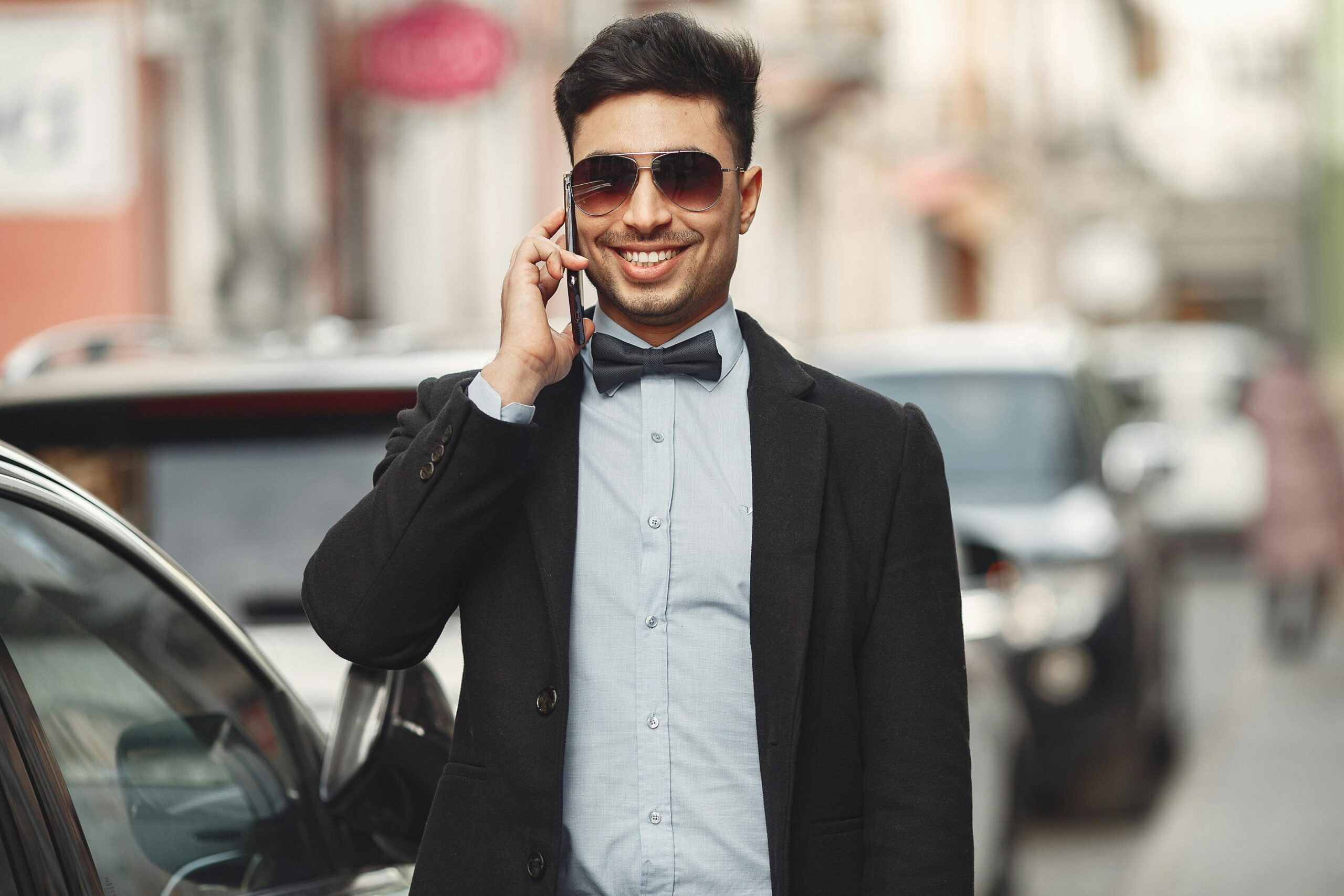 Stylish man wearing sunglasses and a bowtie, making a phone call outdoors.